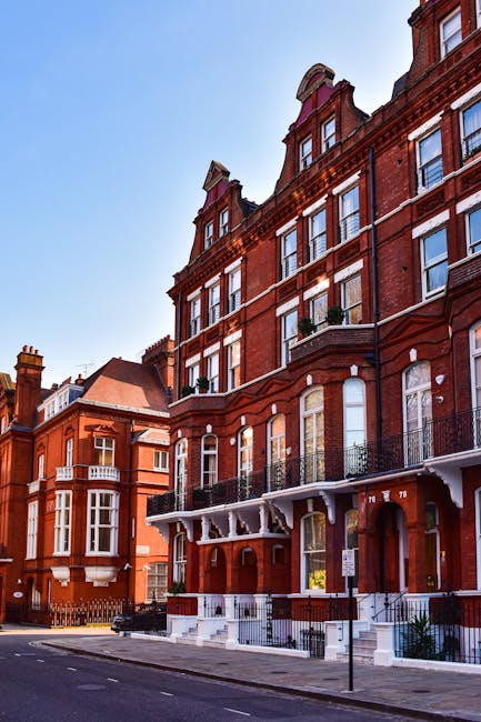 The image depicts a row of historic red-brick Victorian-style terraced houses on a quiet street in Kensington. The buildings feature ornate architectural details, including bay windows with white frames, decorative cornices, and small balconies with wrought-iron railings. The street level shows a well-maintained sidewalk with a few small planters near the entrances. The clear blue sky and soft natural lighting highlight the textures of the red bricks and the details of the facades. The absence of visible garden areas suggests a focus on residential architecture and street scene, typical of the Kensington area. The setting aligns with the themes of property upkeep and exterior appearance, as part of the overall aesthetic associated with the 'Gardeners Kensington' service for local maintenance and landscaping support, as detailed in the 'Gardeners Kensington High Street W8 local maintenance guide' page. This scene emphasizes the charm and elegance of the local neighborhood's exterior environments, with no specific gardening tools or landscape features visible.