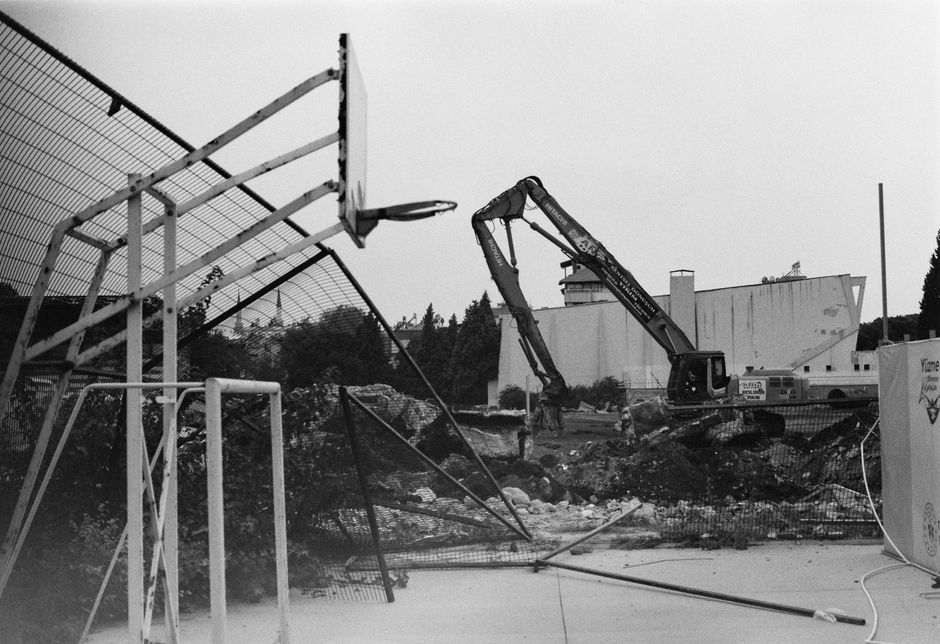 A black and white image showing a construction site with a partially demolished section of a sports court, which includes a basketball hoop and a metal fence. The court appears to be in the process of removal or renovation, with a large excavator working amidst piles of soil, rocks, and debris. In the background, there are industrial-looking buildings and trees, suggesting the area is undergoing landscaping or outdoor space redevelopment. The scene is outdoors with overcast lighting, and the ground shows signs of recent disturbance and excavation, all managed by Gardeners Kensington in line with garden maintenance and landscaping work related to the Gloucester Road to Earls Court gardeners clearance guide.