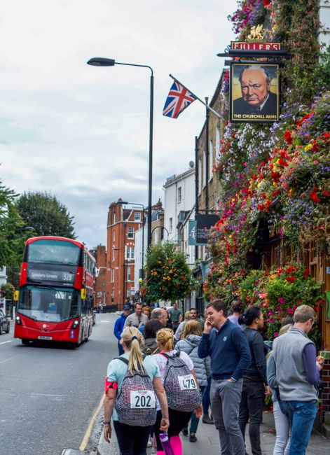 A busy street scene in an urban garden area with a group of people standing on the sidewalk, some wearing numbered bibs suggesting a community walk or event. The sidewalk is lined with vibrant, overflowing flower displays in various colors, including pink, red, purple, and yellow, cascading from hanging baskets and window boxes attached to a building adorned with a pub sign featuring an image of Winston Churchill and a Union Jack flag. In the background, a red double-decker bus is approaching, and the street is flanked by traditional brick and white-painted buildings. The overall environment is lively and well-maintained, with a mix of greenery and urban décor, reflecting garden maintenance and landscaping work in a historic London neighborhood near Gloucester Road to Earls Court. Gardeners Kensington.org.uk might be involved in ensuring the area’s outdoor spaces stay attractive and tidy, complementing the vibrant city life.